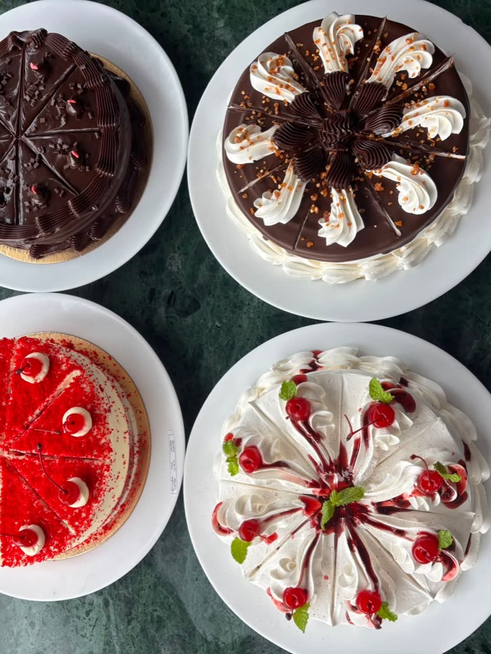 Quatro tortas inteiras sobre a mesa de mármore verde: chocolate, chantilly com cerejas e red velvet